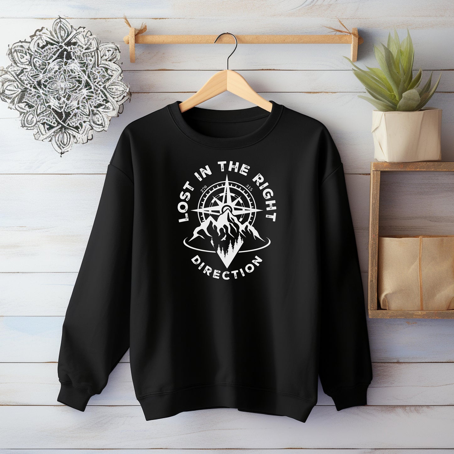 Black sweatshirt featuring a white graphic design of a mountain range with a compass rising behind it, surrounded by pine trees. The words "Lost In The Right Direction" curve around the design, blending adventure and wanderlust vibes. Perfect for outdoor enthusiasts, hikers, and travelers seeking inspiration and style in casual wear.