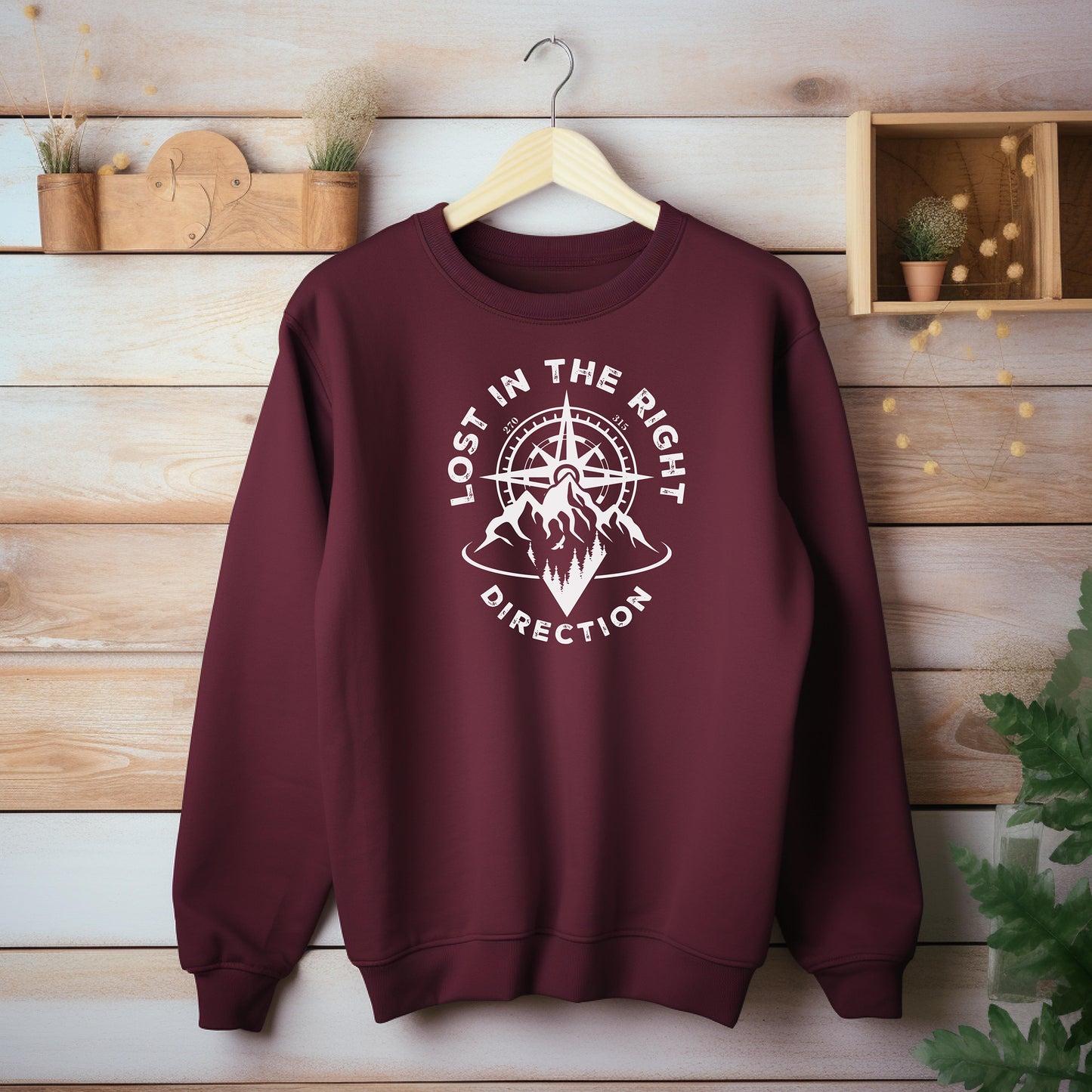 Maroon sweatshirt featuring a white graphic design of a mountain range with a compass rising behind it, surrounded by pine trees. The words "Lost In The Right Direction" curve around the design, blending adventure and wanderlust vibes. Perfect for outdoor enthusiasts, hikers, and travelers seeking inspiration and style in casual wear.