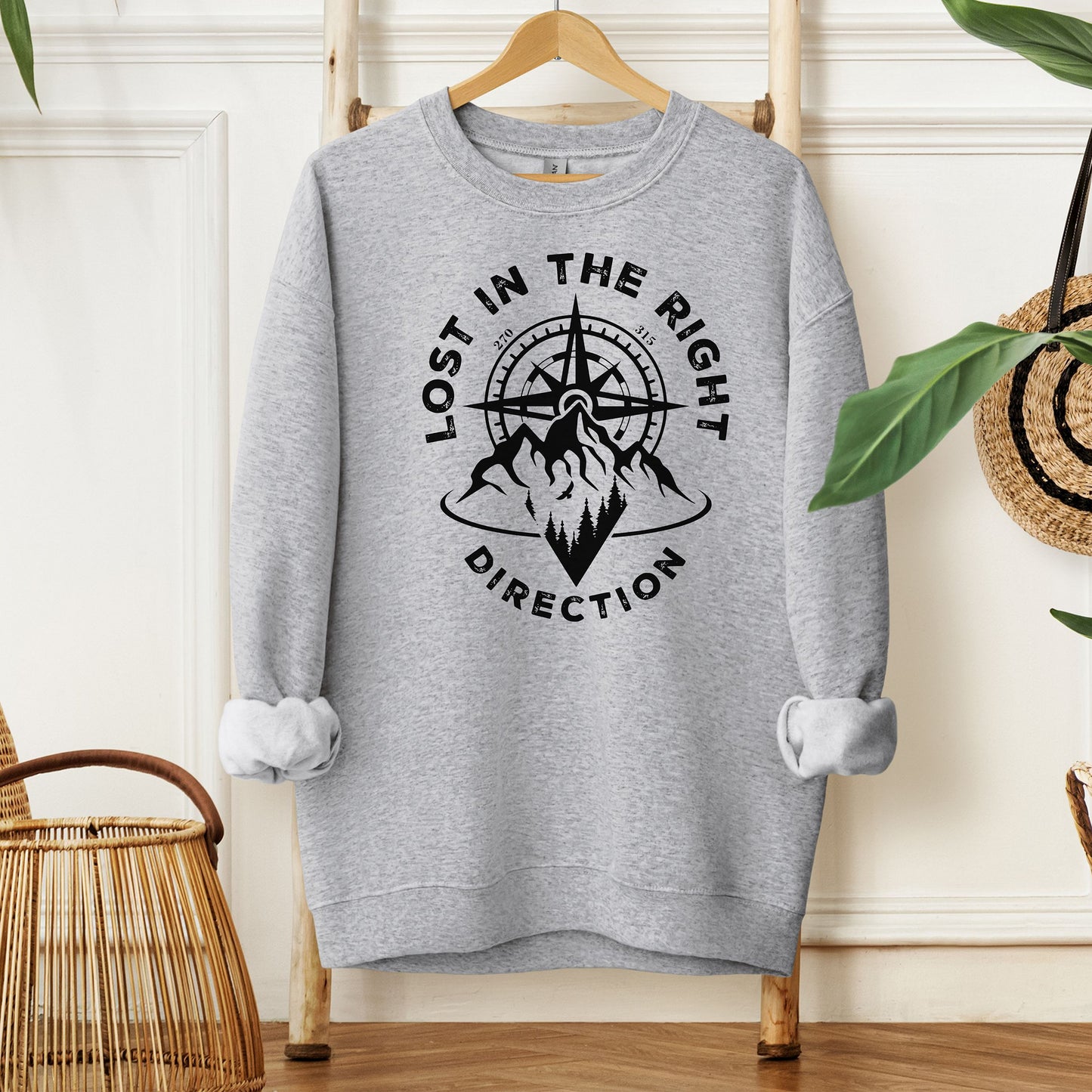 Sport Grey sweatshirt featuring a black graphic design of a mountain range with a compass rising behind it, surrounded by pine trees. The words "Lost In The Right Direction" curve around the design, blending adventure and wanderlust vibes. Perfect for outdoor enthusiasts, hikers, and travelers seeking inspiration and style in casual wear.