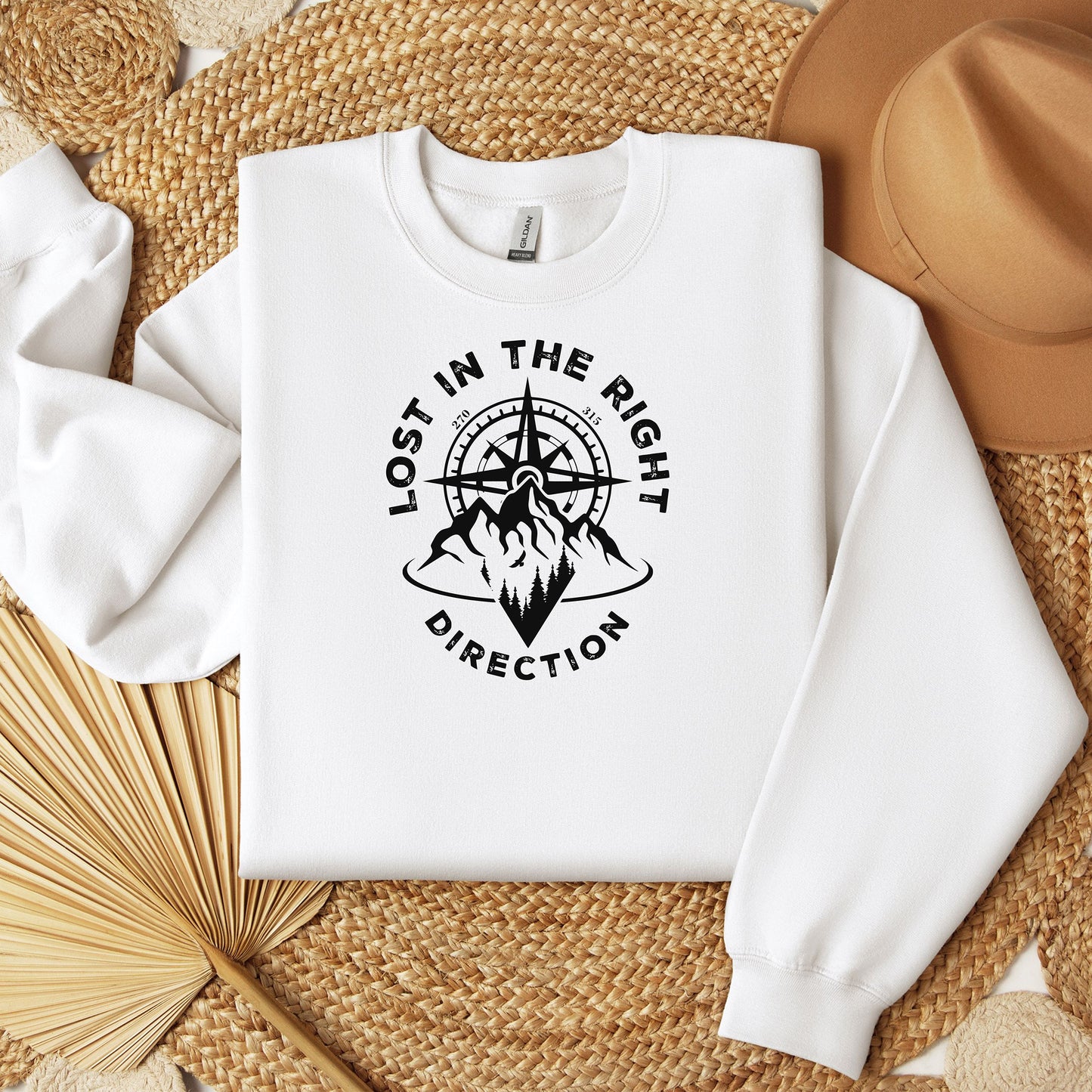 White sweatshirt featuring a black graphic design of a mountain range with a compass rising behind it, surrounded by pine trees. The words "Lost In The Right Direction" curve around the design, blending adventure and wanderlust vibes. Perfect for outdoor enthusiasts, hikers, and travelers seeking inspiration and style in casual wear.