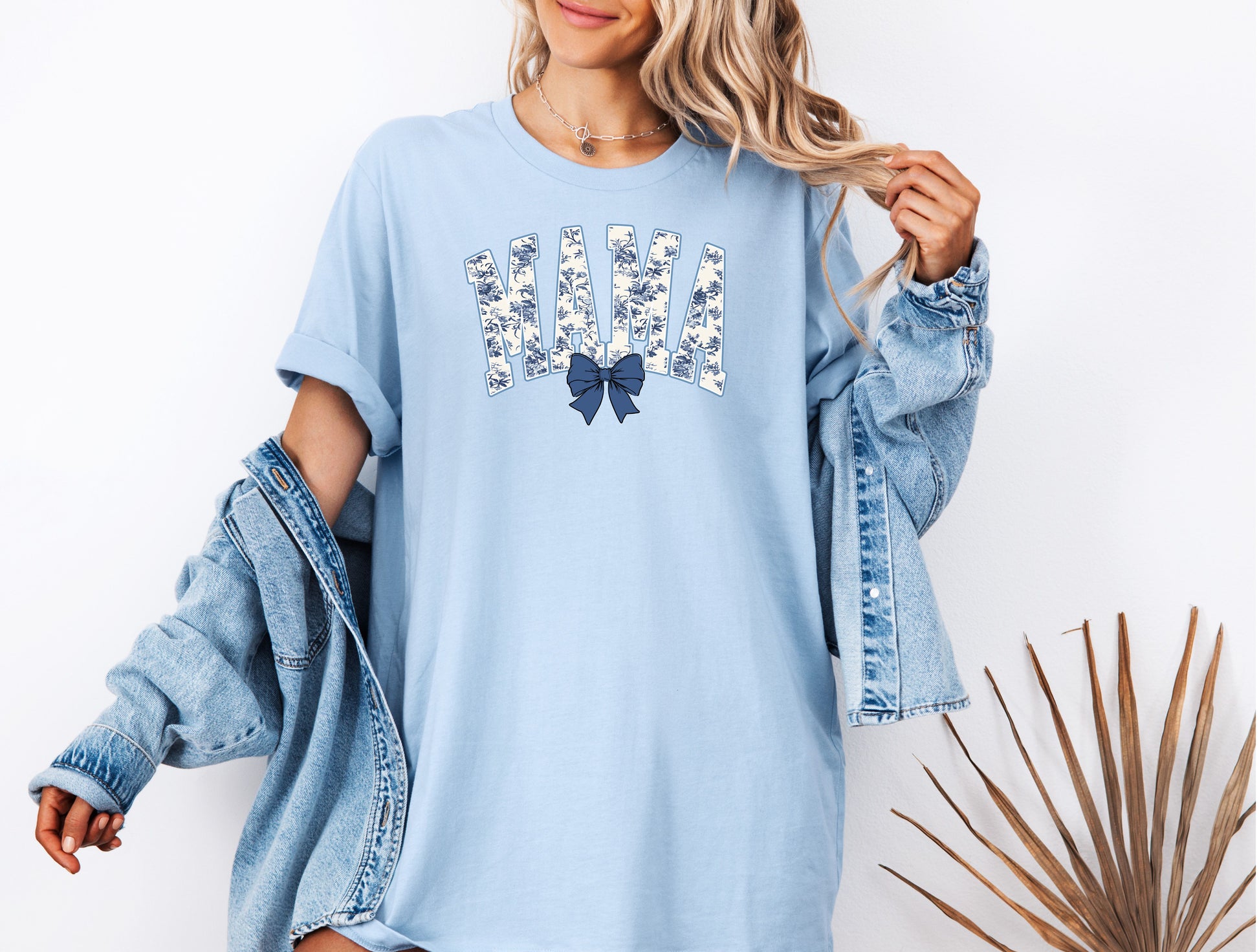 Woman wearing a chambray light blue T-shirt featuring the word “MAMA” in bold block letters filled with a delicate blue toile floral pattern, accented by a matching navy blue coquette bow beneath the text. A stylish and sentimental design perfect for moms, blending classic elegance with casual comfort for everyday wear or gifting.