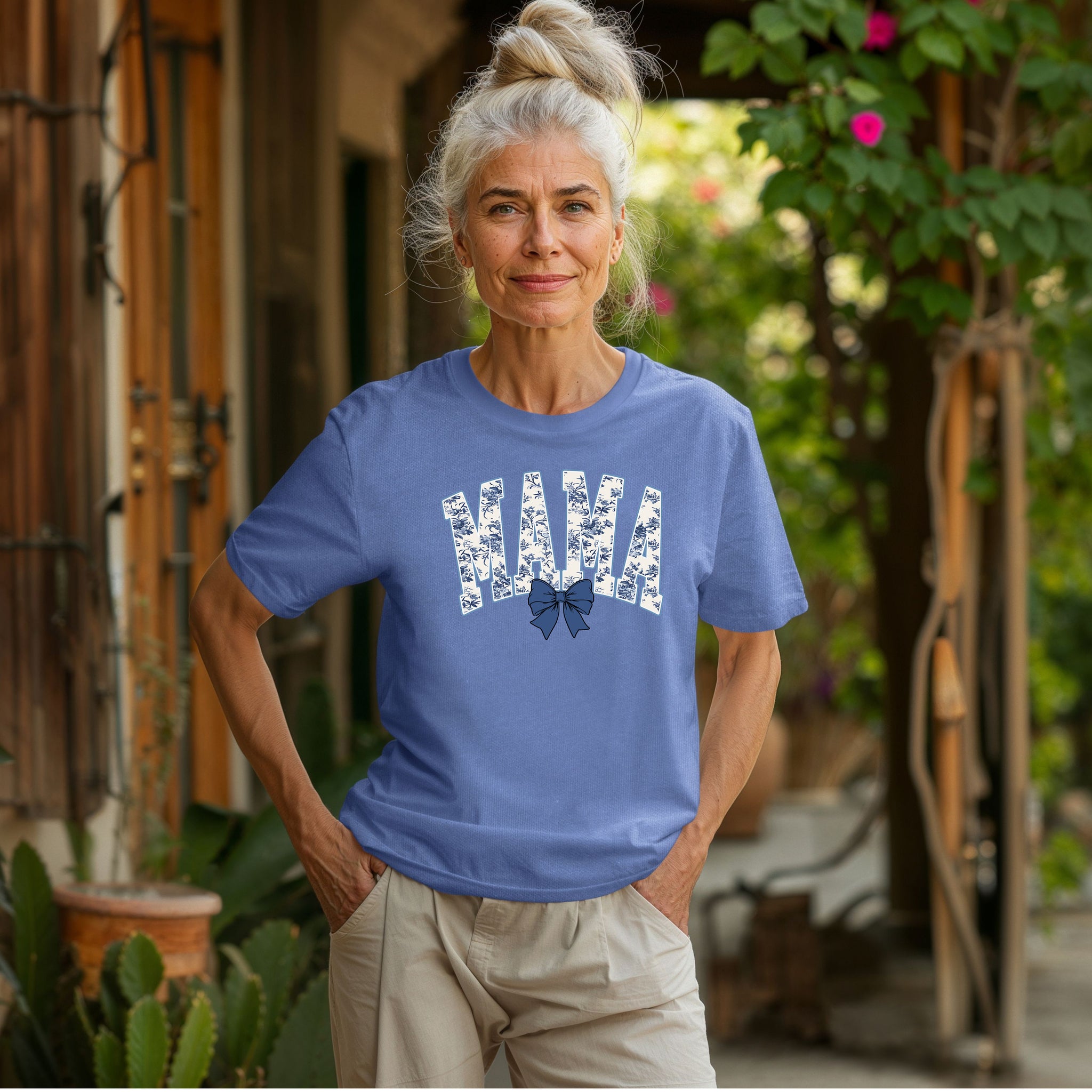 Woman wearing a mystic blue T-shirt featuring the word “MAMA” in bold block letters filled with a delicate blue toile floral pattern, accented by a matching navy blue coquette bow beneath the text. A stylish and sentimental design perfect for moms, blending classic elegance with casual comfort for everyday wear or gifting.