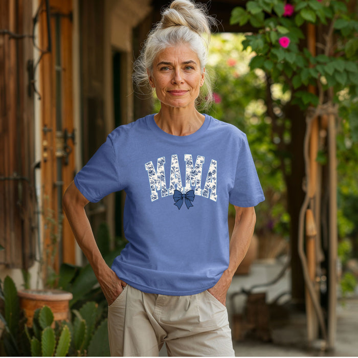 Woman wearing a mystic blue T-shirt featuring the word “MAMA” in bold block letters filled with a delicate blue toile floral pattern, accented by a matching navy blue coquette bow beneath the text. A stylish and sentimental design perfect for moms, blending classic elegance with casual comfort for everyday wear or gifting.
