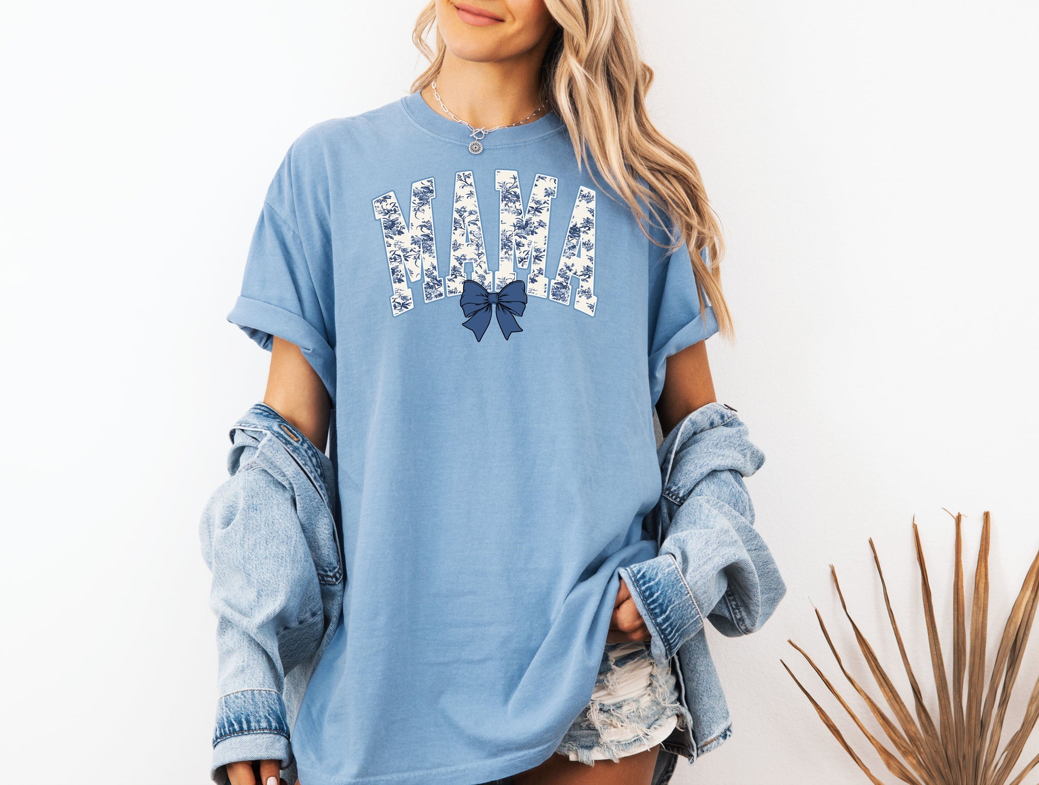 Woman wearing a washed denim T-shirt featuring the word “MAMA” in bold block letters filled with a delicate blue toile floral pattern, accented by a matching navy blue coquette bow beneath the text. A stylish and sentimental design perfect for moms, blending classic elegance with casual comfort for everyday wear or gifting.