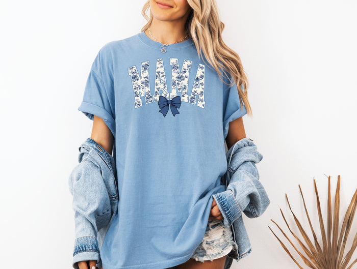 Woman wearing a washed denim T-shirt featuring the word “MAMA” in bold block letters filled with a delicate blue toile floral pattern, accented by a matching navy blue coquette bow beneath the text. A stylish and sentimental design perfect for moms, blending classic elegance with casual comfort for everyday wear or gifting.