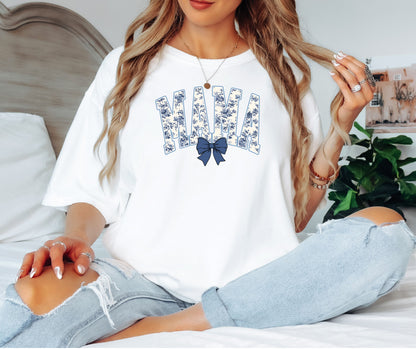 Woman wearing a class white T-shirt featuring the word “MAMA” in bold block letters filled with a delicate blue toile floral pattern, accented by a matching navy blue coquette bow beneath the text. A stylish and sentimental design perfect for moms, blending classic elegance with casual comfort for everyday wear or gifting.