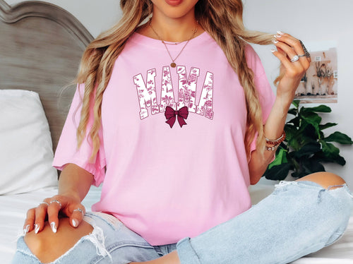 Blossom pink T-shirt featuring a “MAMA” graphic in bold block letters filled with a pink toile floral pattern, accented by a deep maroon bow beneath the text. Styled for a cozy, casual look, this shirt is perfect for moms who love a blend of elegance and comfort, making it an ideal gift for Mother’s Day or everyday wear.