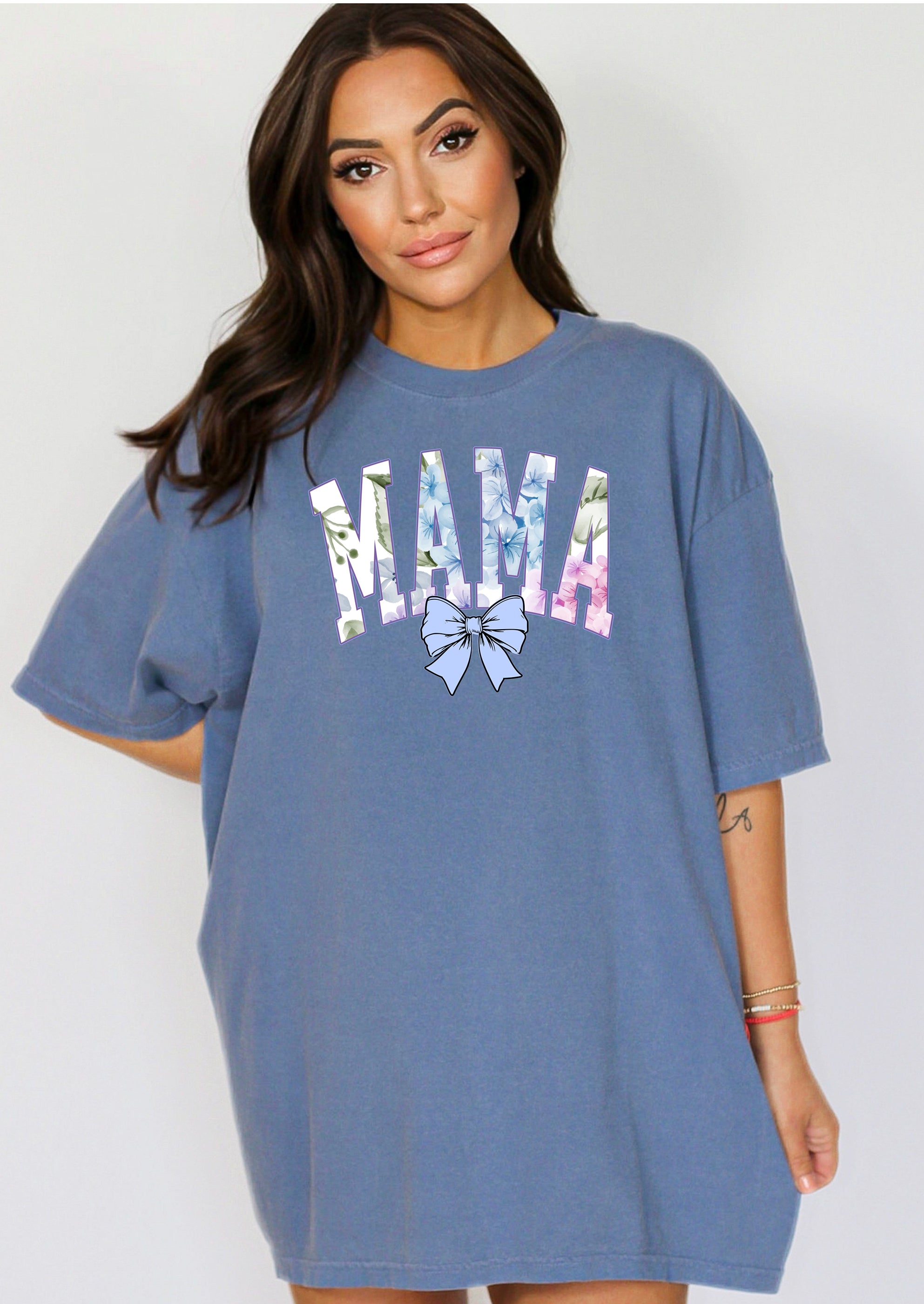 Person wearing a blue jean T-shirt featuring the word “MAMA” in large block letters filled with a delicate blue hydrangea floral pattern, accented with soft green and pink tones. Below the text is a pastel blue coquette bow, adding a sweet, feminine touch.  This spring-inspired graphic tee blends floral elegance with playful charm, perfect for fashionable moms.