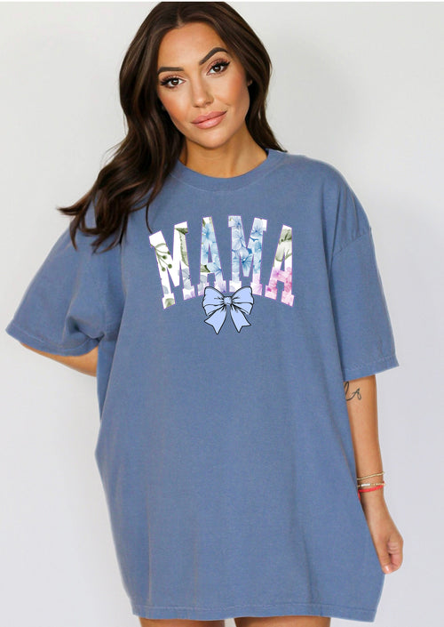 Person wearing a blue jean T-shirt featuring the word “MAMA” in large block letters filled with a delicate blue hydrangea floral pattern, accented with soft green and pink tones. Below the text is a pastel blue coquette bow, adding a sweet, feminine touch.  This spring-inspired graphic tee blends floral elegance with playful charm, perfect for fashionable moms.