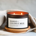 Mango + Coconut Milk candle in a 8 oz black matte candle tin on a wooden coaster with a beige cloth background