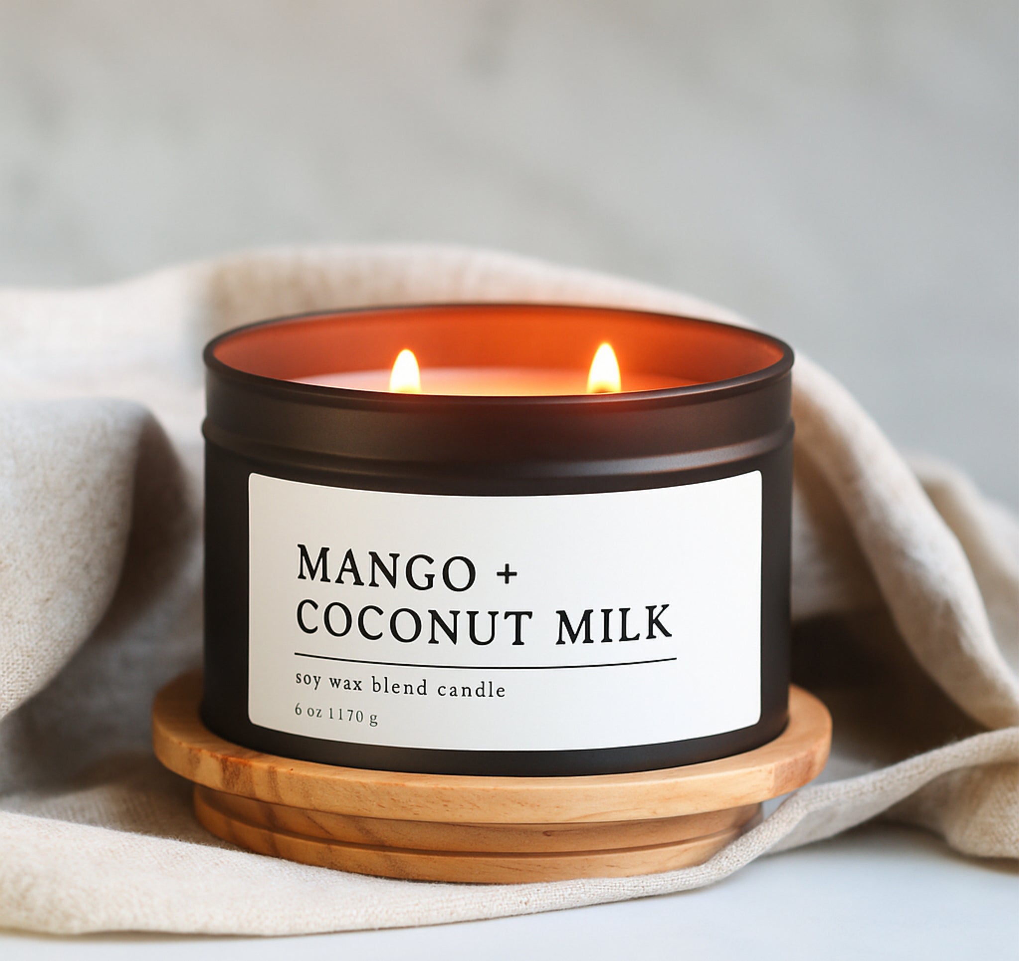 Mango + Coconut Milk candle in a 8 oz black matte candle tin on a wooden coaster with a beige cloth background