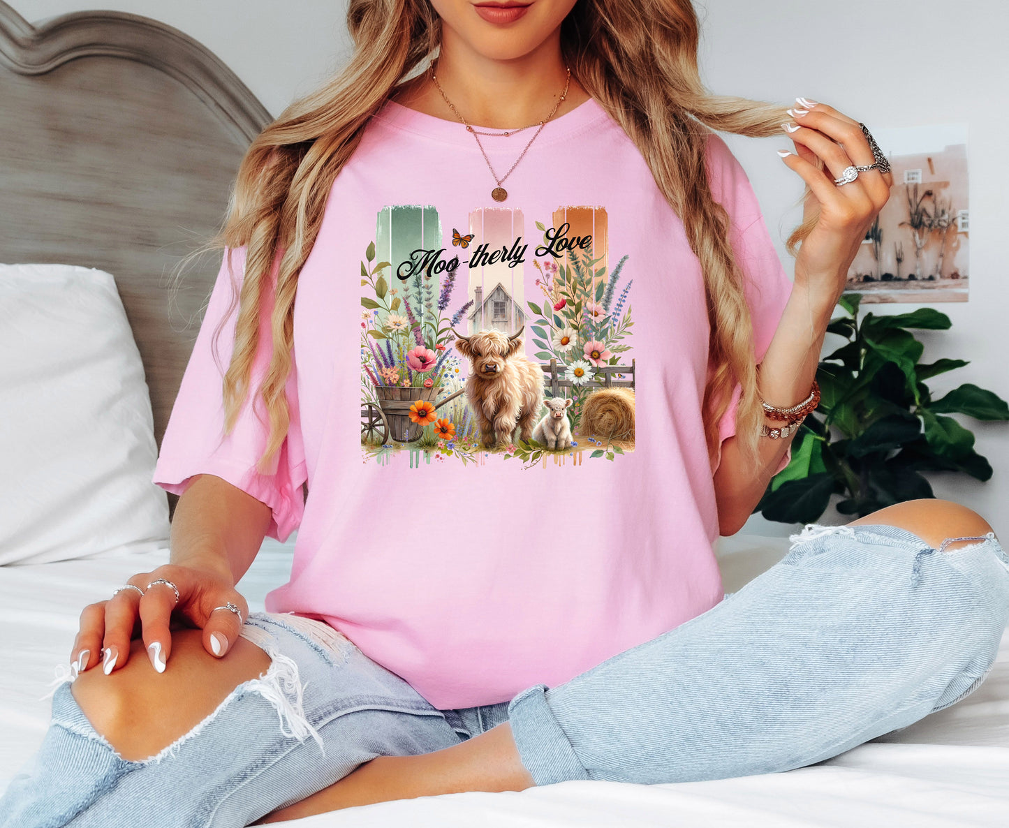 Blossom pink “Moo-therly Love” T-shirt featuring a charming highland cow with her calf, surrounded by colorful wildflowers, greenery, and rustic farm elements. The background showcases three vertical distressed panels in green, cream, and orange, with a quaint barn and butterfly detail. This soft, casual tee blends country charm with sweet, pastoral style, perfect for animal lovers and farm-inspired fashion.