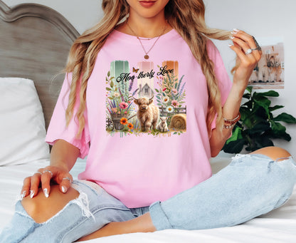 Blossom pink “Moo-therly Love” T-shirt featuring a charming highland cow with her calf, surrounded by colorful wildflowers, greenery, and rustic farm elements. The background showcases three vertical distressed panels in green, cream, and orange, with a quaint barn and butterfly detail. This soft, casual tee blends country charm with sweet, pastoral style, perfect for animal lovers and farm-inspired fashion.