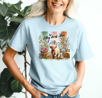 Chambray light blue “Moo-therly Love” T-shirt featuring a charming highland cow with her calf, surrounded by colorful wildflowers, greenery, and rustic farm elements. The background showcases three vertical distressed panels in green, cream, and orange, with a quaint barn and butterfly detail. This soft, casual tee blends country charm with sweet, pastoral style, perfect for animal lovers and farm-inspired fashion.
