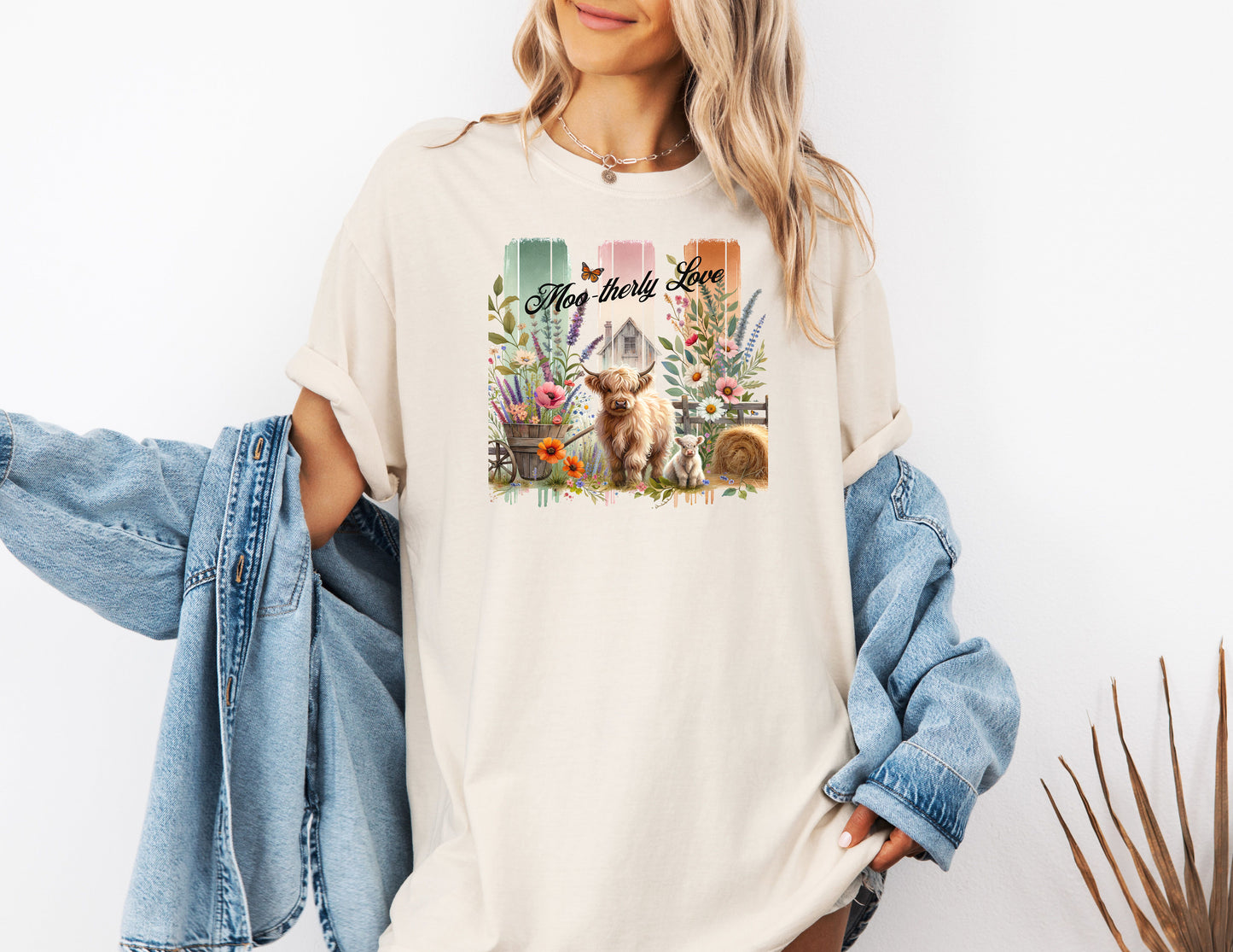 Ivory “Moo-therly Love” T-shirt featuring a charming highland cow with her calf, surrounded by colorful wildflowers, greenery, and rustic farm elements. The background showcases three vertical distressed panels in green, cream, and orange, with a quaint barn and butterfly detail. This soft, casual tee blends country charm with sweet, pastoral style, perfect for animal lovers and farm-inspired fashion.