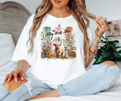White “Moo-therly Love” T-shirt featuring a charming highland cow with her calf, surrounded by colorful wildflowers, greenery, and rustic farm elements. The background showcases three vertical distressed panels in green, cream, and orange, with a quaint barn and butterfly detail. This soft, casual tee blends country charm with sweet, pastoral style, perfect for animal lovers and farm-inspired fashion.