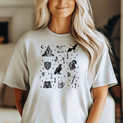 Person wearing an Ash T-shirt featuring the "Moonlit Magick" Renaissance Faire design. The shirt showcases a collection of detailed black illustrations, including a medieval castle, a dragon, knight helmet, shield, raven, spellbook, sword, key, mug of ale, and stars—capturing the enchanting spirit of medieval fantasy.