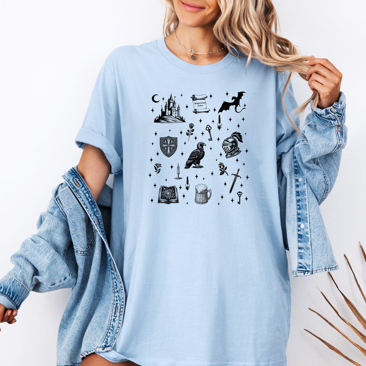 Person wearing a baby blue T-shirt featuring the "Moonlit Magick" Renaissance Faire design. The shirt showcases a collection of detailed black illustrations, including a medieval castle, a dragon, knight helmet, shield, raven, spellbook, sword, key, mug of ale, and stars—capturing the enchanting spirit of medieval fantasy.