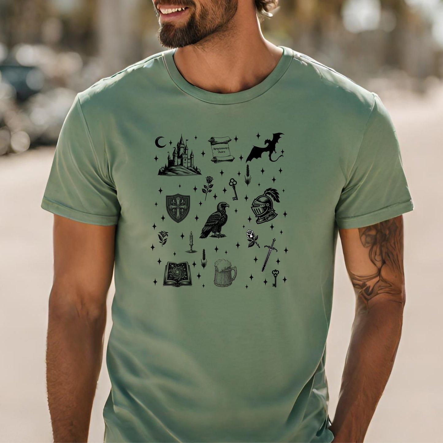 Man wearing a Sage T-shirt featuring the "Moonlit Magick" Renaissance Faire design. The shirt showcases a collection of detailed black illustrations, including a medieval castle, a dragon, knight helmet, shield, raven, spellbook, sword, key, mug of ale, and stars—capturing the enchanting spirit of medieval fantasy.