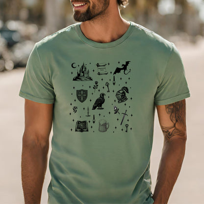 Man wearing a Sage T-shirt featuring the "Moonlit Magick" Renaissance Faire design. The shirt showcases a collection of detailed black illustrations, including a medieval castle, a dragon, knight helmet, shield, raven, spellbook, sword, key, mug of ale, and stars—capturing the enchanting spirit of medieval fantasy.