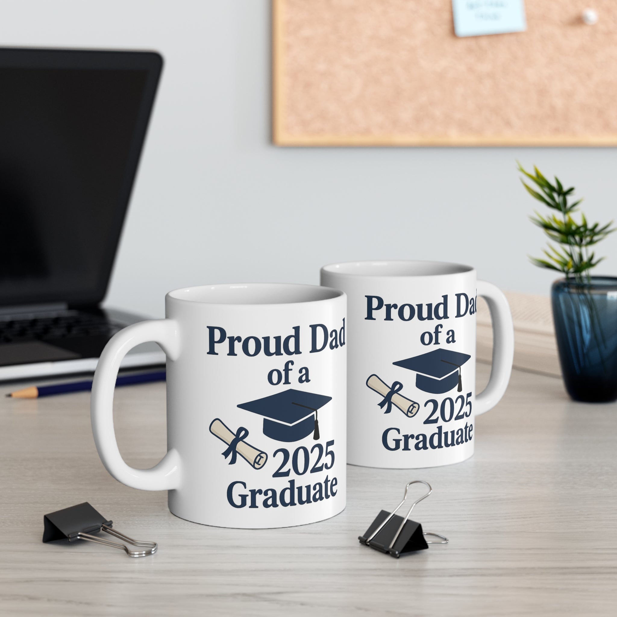 Two mugs with 'Proud Dad of a 2025 Graduate' text, a diploma, and a graduation cap sitting on a desk with office supplies.