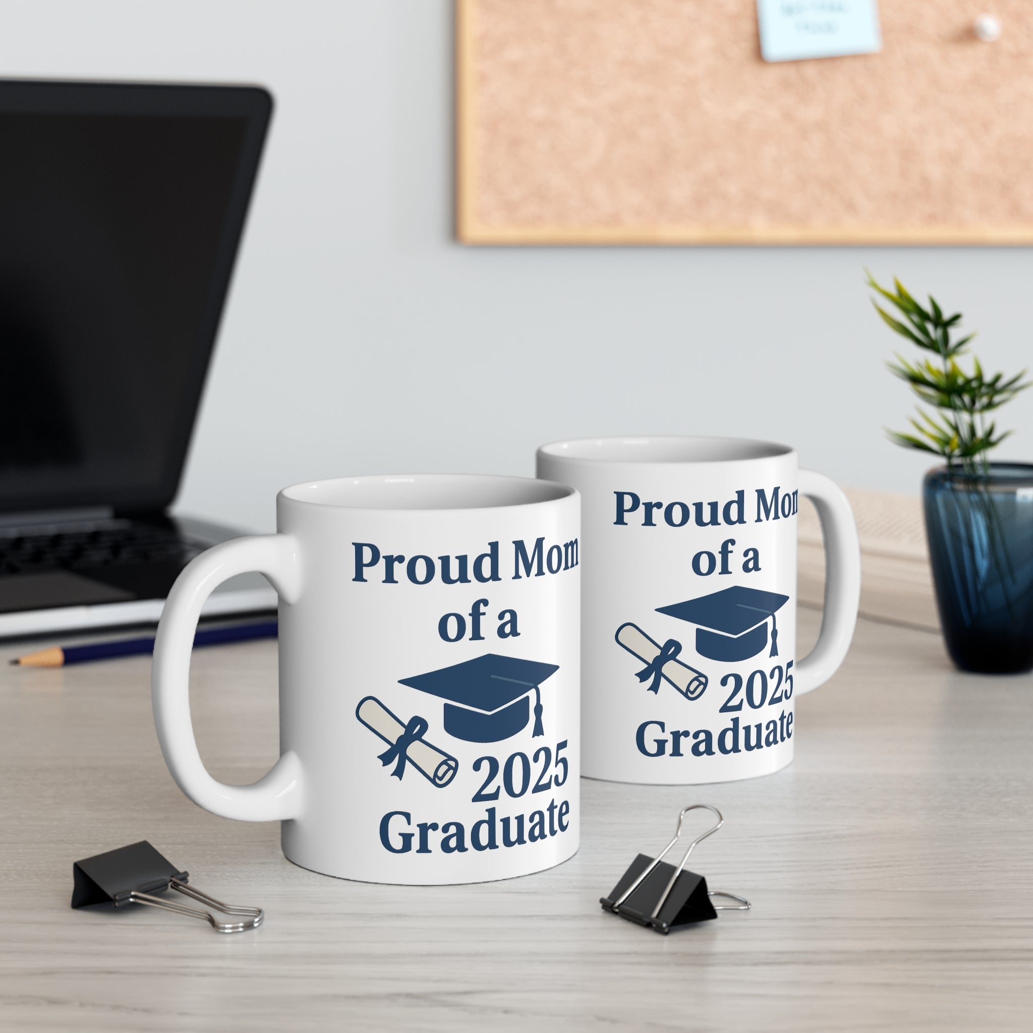Two white mugs with 'Proud Mom of a 2025 Graduate' text, a diploma, and a graduation cap sitting on a desk with office supplies.