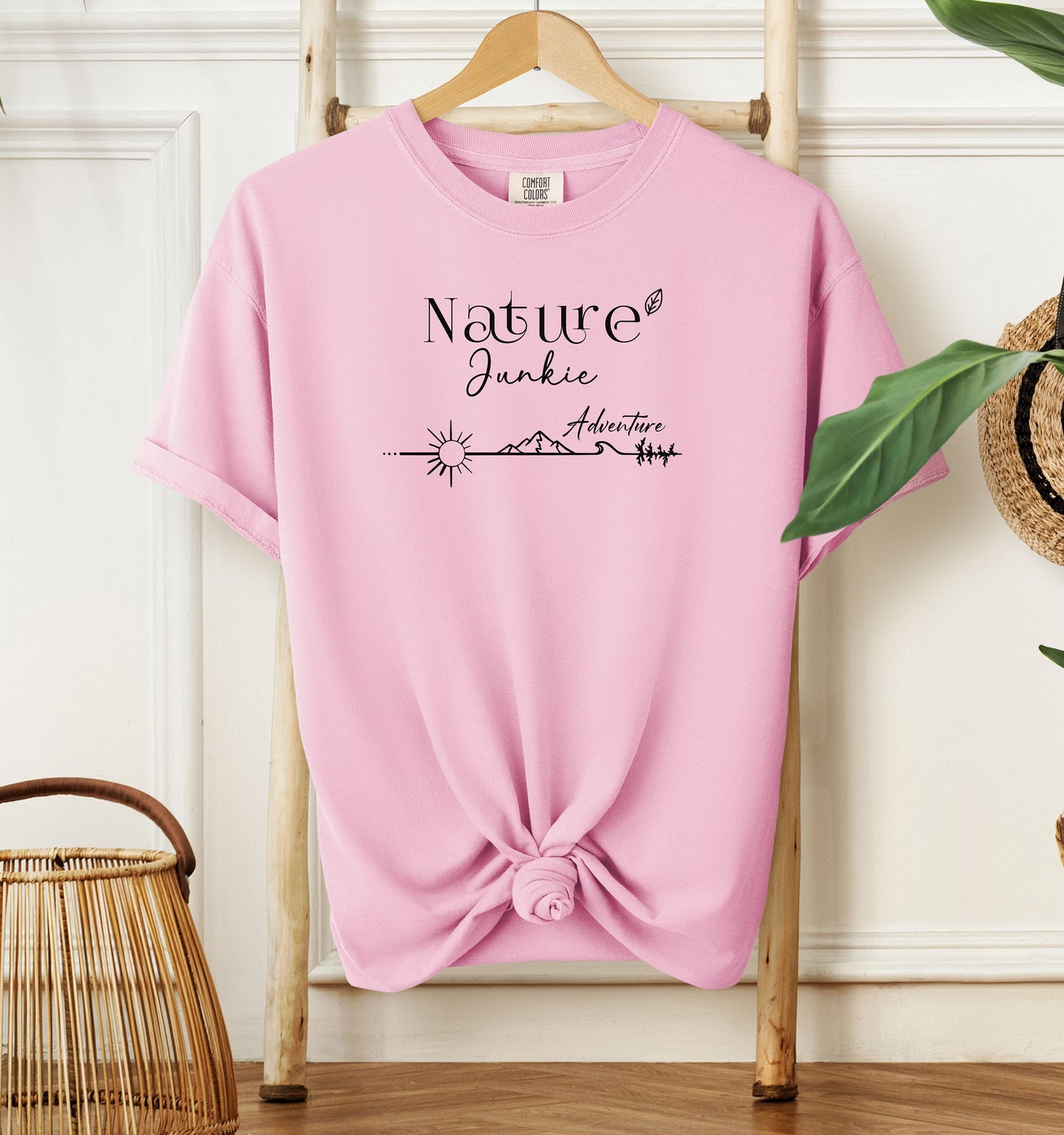 Nature Junkie T-shirt with minimalist black line art showing a sun, mountains, trees, and the words 'Nature Junkie Adventure' with a small leaf accent. Printed on a soft blossom pink unisex Comfort Colors tee, perfect for outdoor lovers, hikers, campers, and those who enjoy exploring nature and adventure.