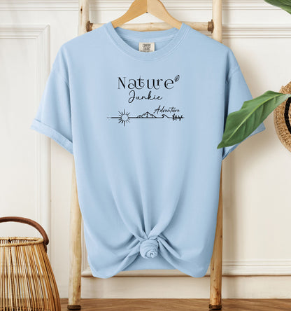 Nature Junkie T-shirt with minimalist black line art showing a sun, mountains, trees, and the words 'Nature Junkie Adventure' with a small leaf accent. Printed on a soft chambray light blue unisex Comfort Colors tee, perfect for outdoor lovers, hikers, campers, and those who enjoy exploring nature and adventure.