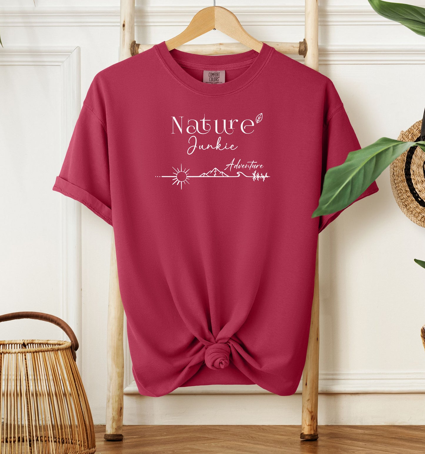 Nature Junkie T-shirt with minimalist white line art showing a sun, mountains, trees, and the words 'Nature Junkie Adventure' with a small leaf accent. Printed on a soft chili red unisex Comfort Colors tee, perfect for outdoor lovers, hikers, campers, and those who enjoy exploring nature and adventure.