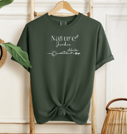 Nature Junkie T-shirt with minimalist white line art showing a sun, mountains, trees, and the words 'Nature Junkie Adventure' with a small leaf accent. Printed on a soft Hemp color unisex Comfort Colors tee, perfect for outdoor lovers, hikers, campers, and those who enjoy exploring nature and adventure.
