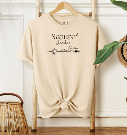 Nature Junkie T-shirt with minimalist black line art showing a sun, mountains, trees, and the words 'Nature Junkie Adventure' with a small leaf accent. Printed on a soft ivory unisex Comfort Colors tee, perfect for outdoor lovers, hikers, campers, and those who enjoy exploring nature and adventure.
