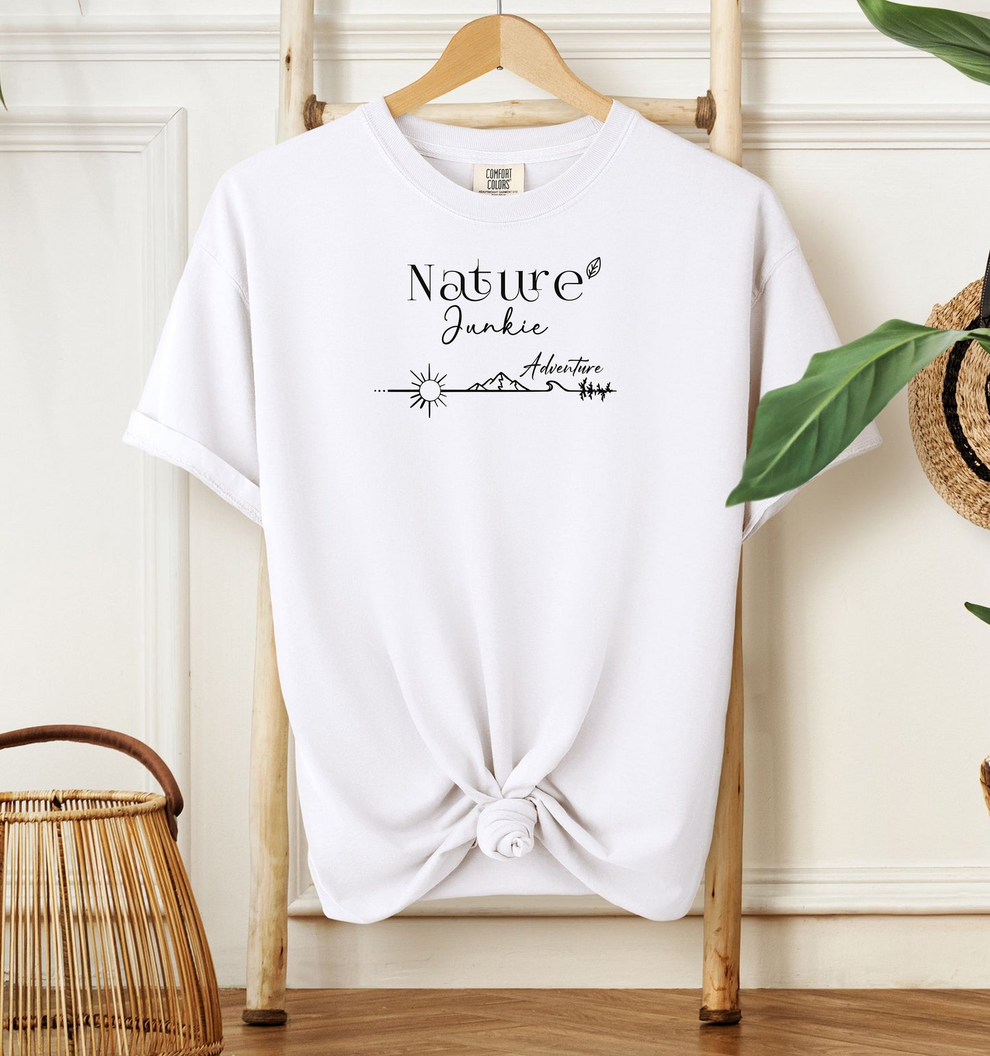 Nature Junkie T-shirt with minimalist black line art showing a sun, mountains, trees, and the words 'Nature Junkie Adventure' with a small leaf accent. Printed on a soft white unisex Comfort Colors tee, perfect for outdoor lovers, hikers, campers, and those who enjoy exploring nature and adventure.