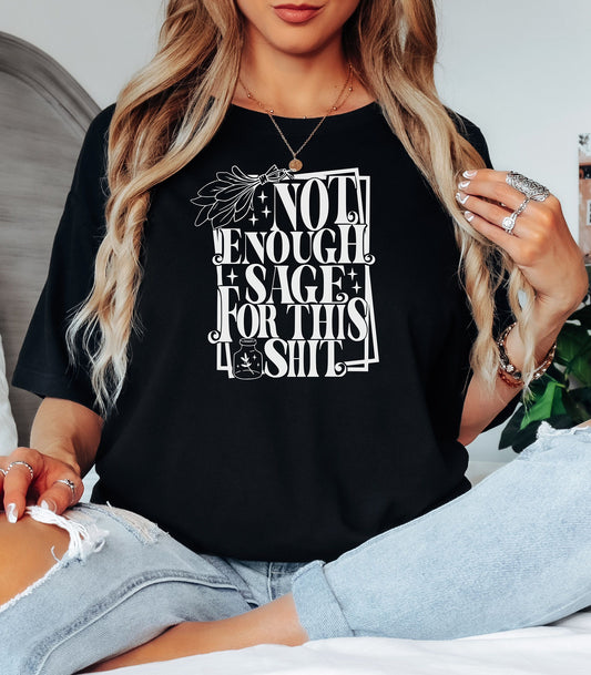 Black T-shirt with bold white typography reading “Not Enough Sage For This Shit,” accented with illustrated sage leaves, sparkles, and a small potion jar with a leafy design. Playful and cheeky statement shirt for spiritual, witchy, or humor-loving personalities.