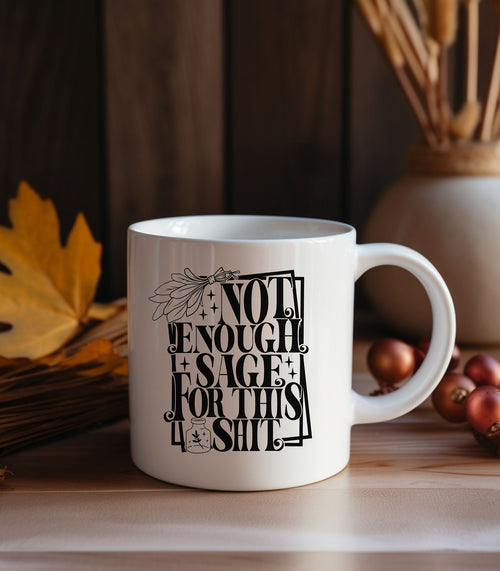 White ceramic mug featuring bold black typography reading “Not Enough Sage For This Shit,” framed in a whimsical outlined border. The design includes a detailed sage bundle illustration at the top and a small jar with a sage leaf motif at the bottom, adding a witchy, mystical vibe. Perfect for humorous coffee or tea moments, it blends sassy attitude with boho and spiritual flair, making it an ideal gift for friends with a sense of humor.
