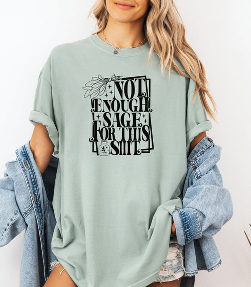 Bay light green T-shirt with bold black typography reading “Not Enough Sage For This Shit,” accented with illustrated sage leaves, sparkles, and a small potion jar with a leafy design. Playful and cheeky statement shirt for spiritual, witchy, or humor-loving personalities.