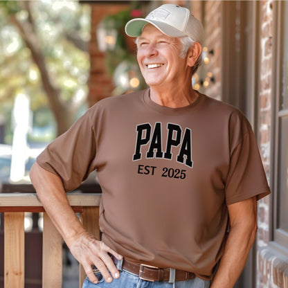 PAPA Est [Year] T-shirt featuring bold collegiate-style lettering with a customizable establishment year. Printed on a soft, relaxed-fit unisex tee in a espresso color, this design makes a perfect gift for new grandfathers, Father’s Day, or family celebrations. Ideal for proudly announcing the year someone became 'Papa' while offering comfort and casual style.