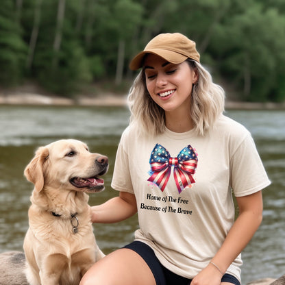 Ivory t-shirt featuring a patriotic coquette bow design with stars and stripes in red, white, and blue. Below the bow, the text reads 'Home of The Free Because of The Brave.' A stylish and feminine Holiday shirt, perfect for celebrating Independence Day, parades, and summer festivities.