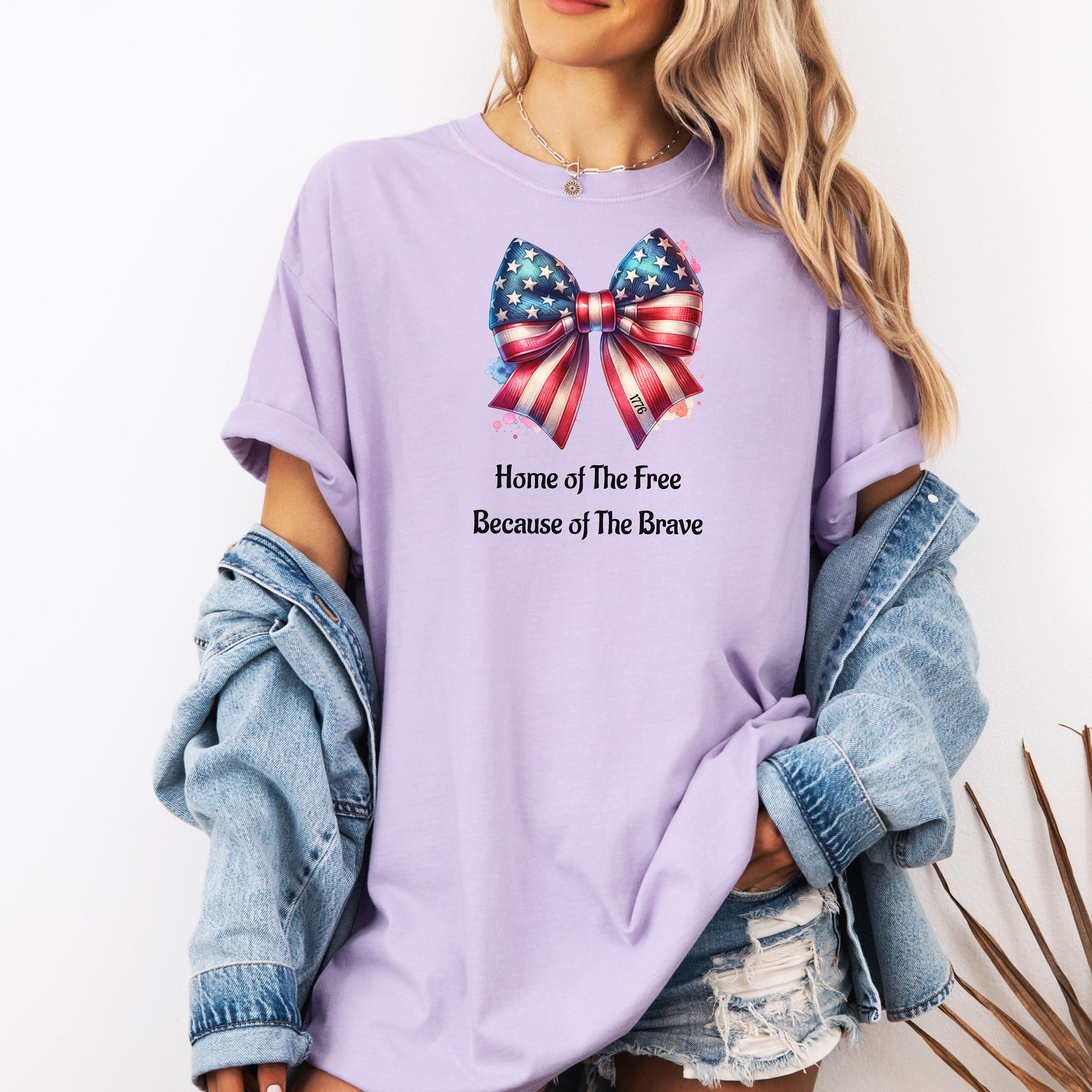 Orchid t-shirt featuring a patriotic coquette bow design with stars and stripes in red, white, and blue. Below the bow, the text reads 'Home of The Free Because of The Brave.' A stylish and feminine Holiday shirt, perfect for celebrating Independence Day, parades, and summer festivities.