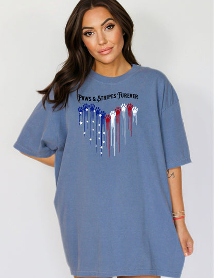 Person wearing a blue jean T-shirt featuring a heart-shaped design made of dripping red, white, and blue paw prints with star accents. Above the heart, the text reads “Paws & Stripes Furever,” blending patriotic pride with a love for pets, making it a perfect shirt for animal lovers and USA enthusiasts.