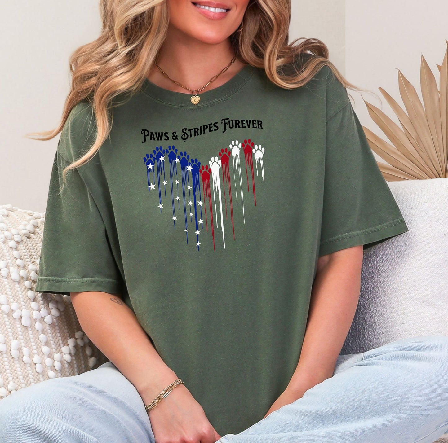 Person wearing a hemp color T-shirt featuring a heart-shaped design made of dripping red, white, and blue paw prints with star accents. Above the heart, the text reads “Paws & Stripes Furever,” blending patriotic pride with a love for pets, making it a perfect shirt for animal lovers and USA enthusiasts.