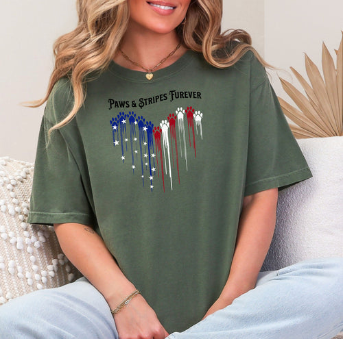 Person wearing a hemp color T-shirt featuring a heart-shaped design made of dripping red, white, and blue paw prints with star accents. Above the heart, the text reads “Paws & Stripes Furever,” blending patriotic pride with a love for pets, making it a perfect shirt for animal lovers and USA enthusiasts.