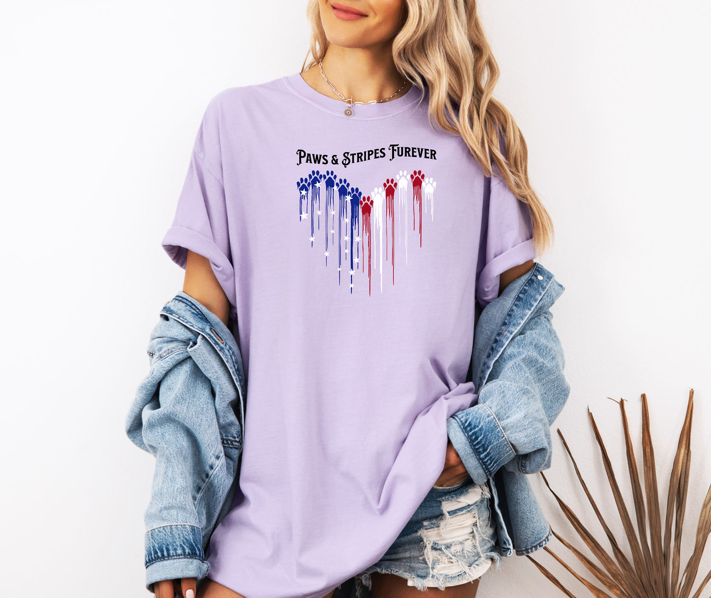 Person wearing an orchid T-shirt featuring a heart-shaped design made of dripping red, white, and blue paw prints with star accents. Above the heart, the text reads “Paws & Stripes Furever,” blending patriotic pride with a love for pets, making it a perfect shirt for animal lovers and USA enthusiasts.