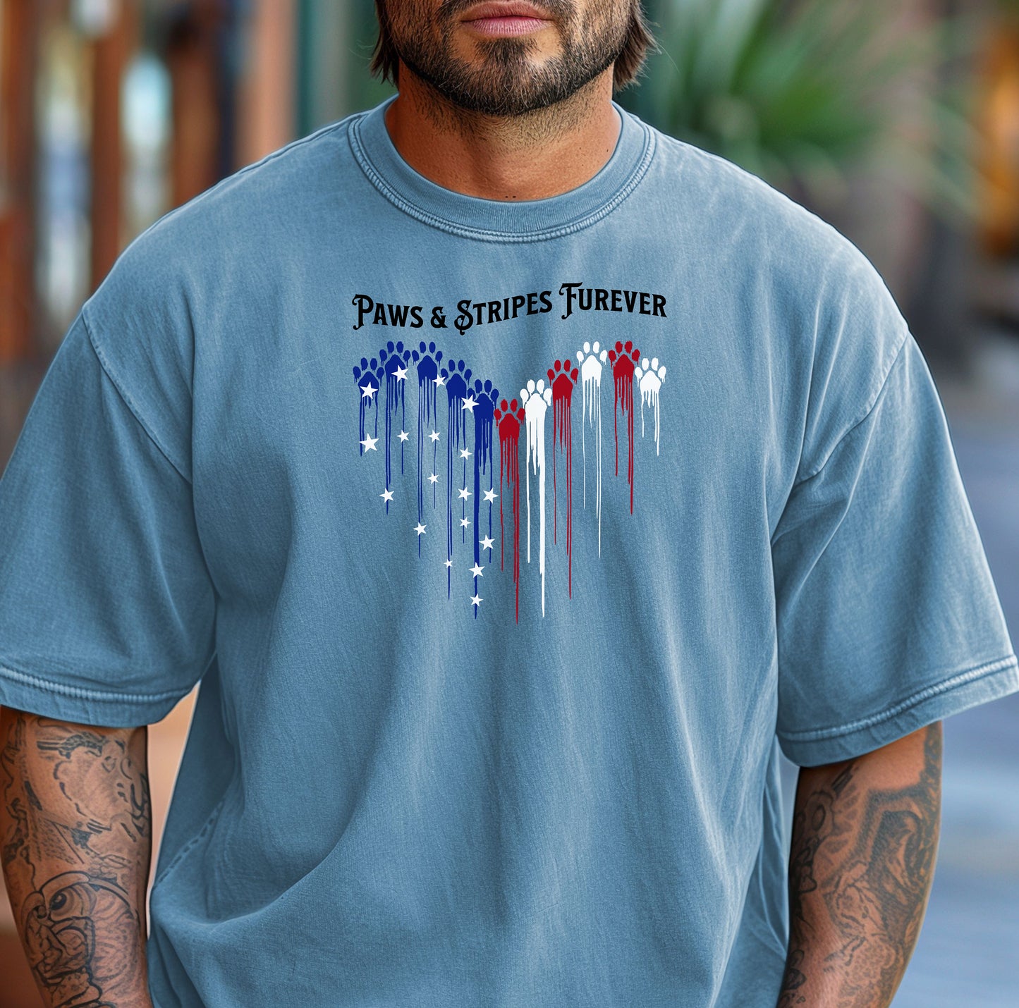 Person wearing a washed denim T-shirt featuring a heart-shaped design made of dripping red, white, and blue paw prints with star accents. Above the heart, the text reads “Paws & Stripes Furever,” blending patriotic pride with a love for pets, making it a perfect shirt for animal lovers and USA enthusiasts.