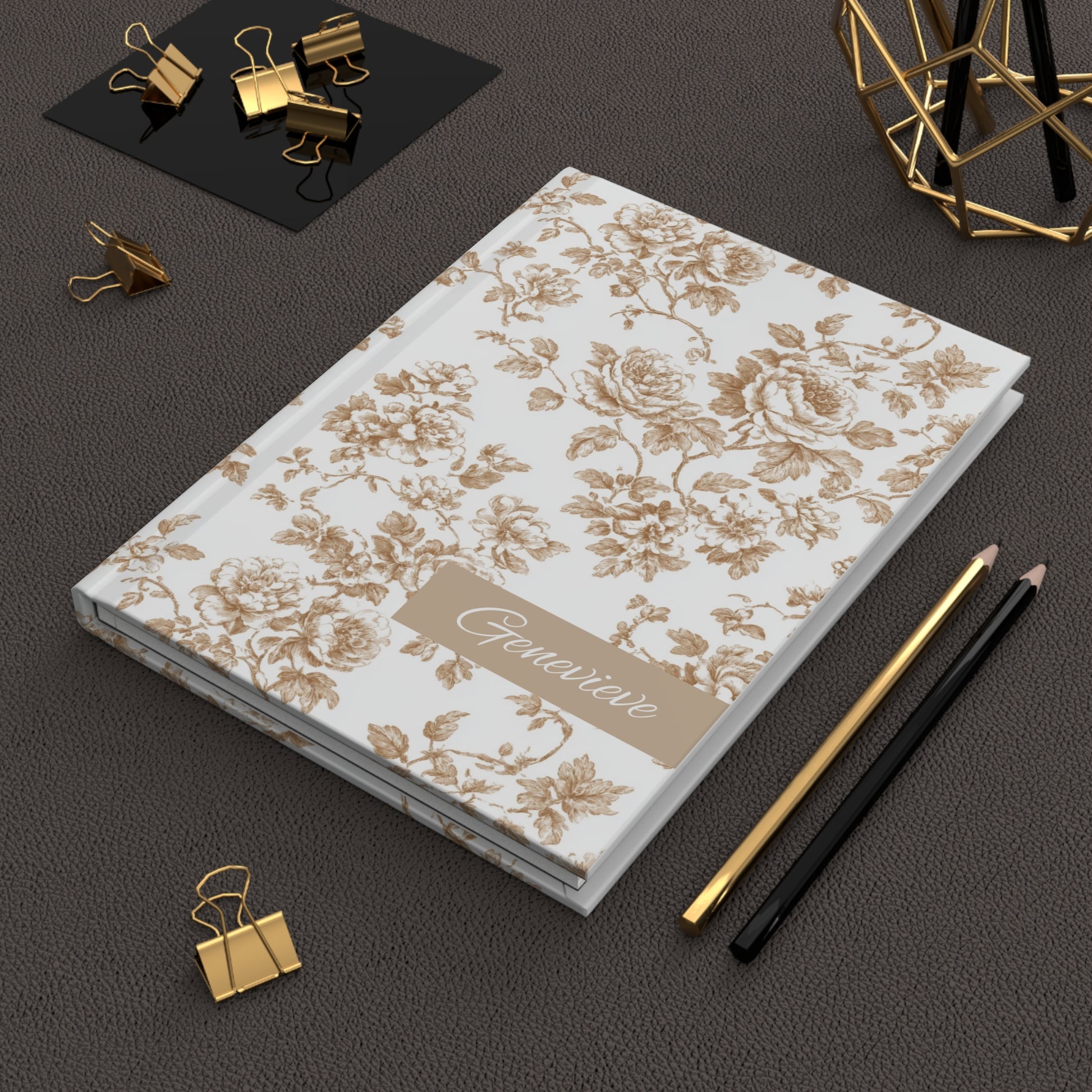 Champagne floral toile patterned hardcover journal with 'Genevieve' on a lying on dark surface with office supplies.