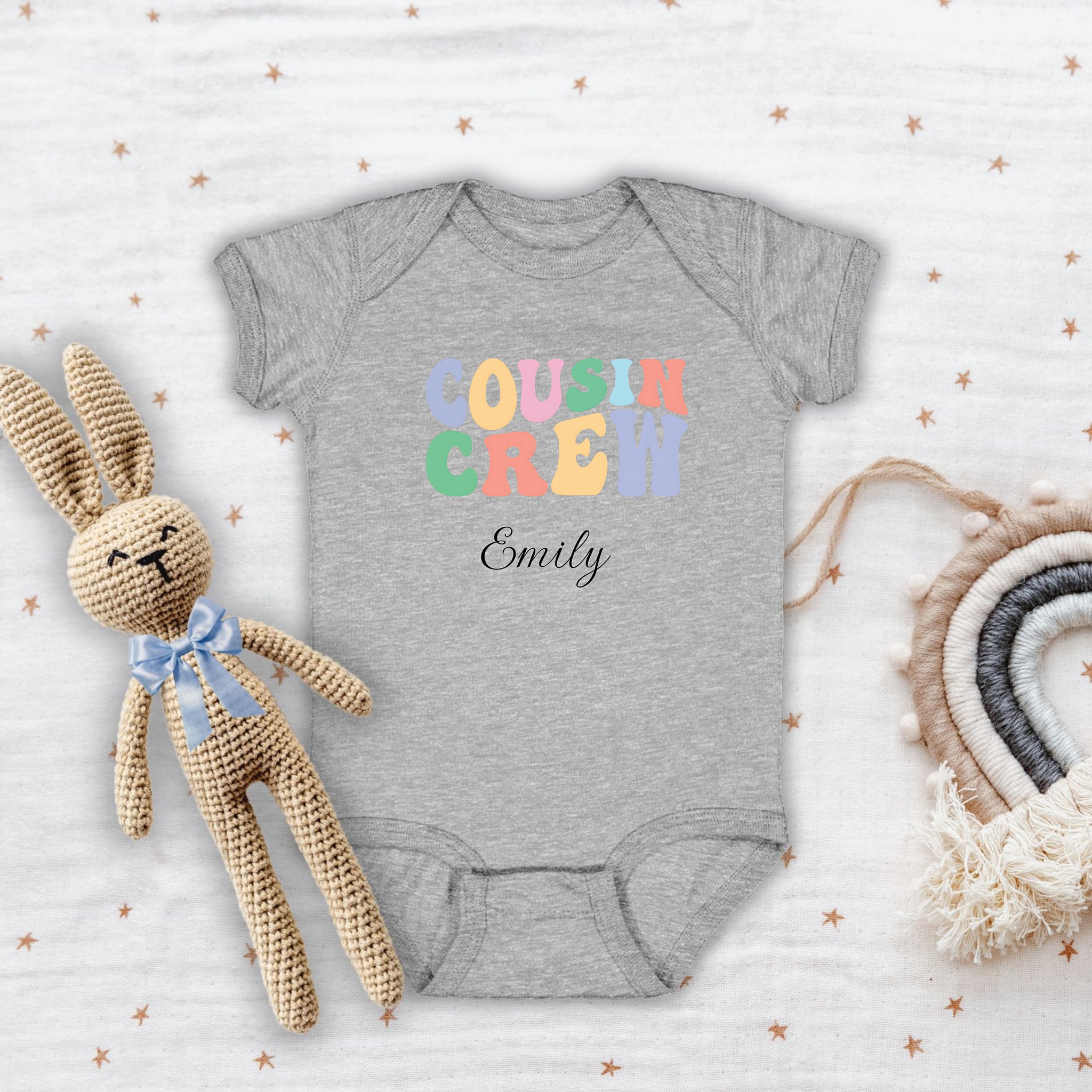 Heather infant bodysuit laid flat on a wooden surface, featuring a colorful pastel “Cousin Crew” design with the name “Your Name” in elegant black script below. Surrounded by cozy knitted bunny and a rainbow mobile.