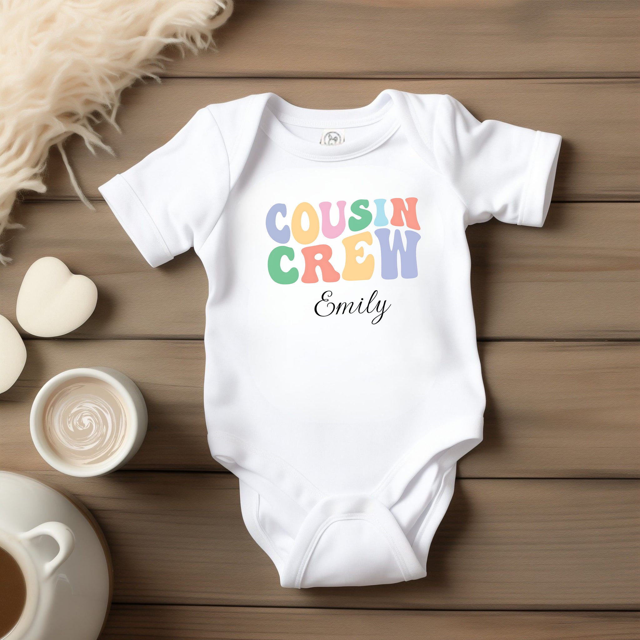 White infant bodysuit laid flat on a wooden surface, featuring a colorful pastel “Cousin Crew” design with the name “Your Name” in elegant black script below. Surrounded by cozy props including a heart-shaped soap, and a fluffy cream-colored blanket for a warm, welcoming feel.