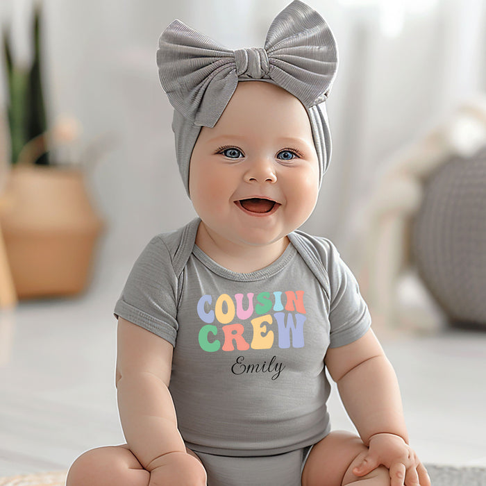 Smiling baby wearing a Heather infant bodysuit featuring a colorful “Cousin Crew” design in playful pastel block letters, with the name “Your Name” written in black script below. Seated on a soft surface next to a plush teddy bear, surrounded by cozy, light-filled decor and greenery in the background.