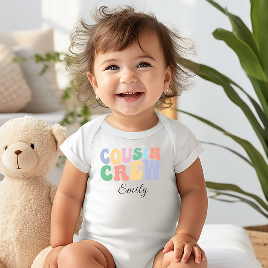 Smiling baby wearing a white infant bodysuit featuring a colorful “Cousin Crew” design in playful pastel block letters, with the name “Your Name” written in black script below. Seated on a soft surface next to a plush teddy bear, surrounded by cozy, light-filled decor and greenery in the background.