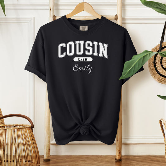 Black Comfort Colors T-shirt featuring a bold “Cousin Crew” design in white varsity-style lettering, with the name “Your Name” in elegant script below. Displayed knotted at the bottom and hanging on a wooden hanger in a cozy, neutral-toned room with natural decor elements.