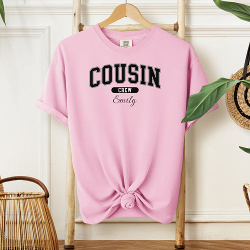 Blossom pink Comfort Colors T-shirt featuring a bold “Cousin Crew” design in black varsity-style lettering, with the name “Your Name” in elegant script below. Displayed knotted at the bottom and hanging on a wooden hanger in a cozy, neutral-toned room with natural decor elements.